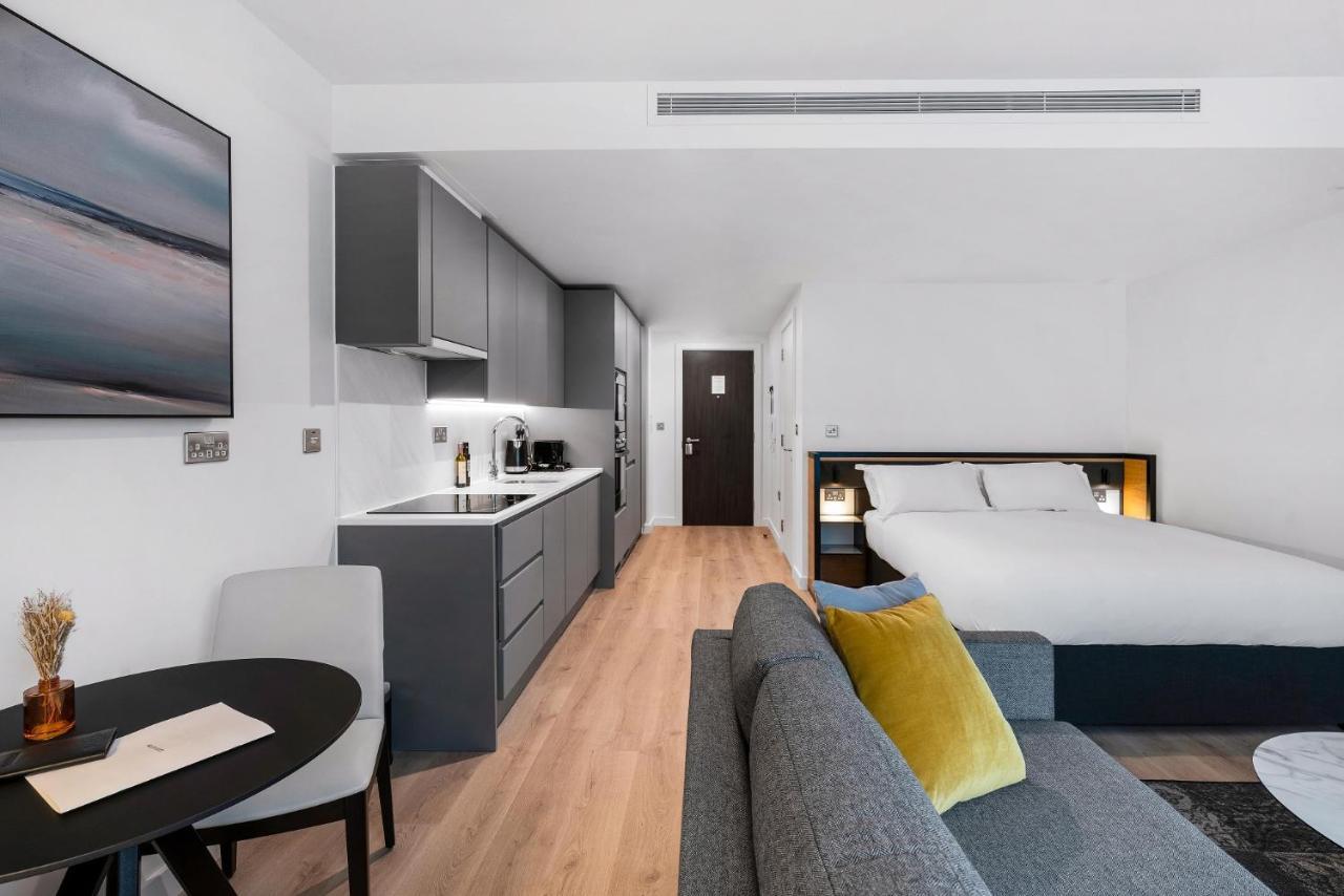 מנצ'סטר Citysuites 2 Aparthotel מראה חיצוני תמונה The photo shows a modern and stylish studio apartment. It features a kitchenette with dark cabinetry and a small dining table with two chairs. There's a cozy sofa with colorful cushions, positioned next to the table. In the background, a large bed wi