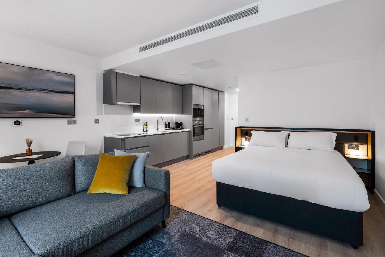 מנצ'סטר Citysuites 2 Aparthotel מראה חיצוני תמונה The photo shows a modern studio apartment. On one side, there is a comfortable gray sofa adorned with a yellow cushion. Nearby, a round table is placed, likely intended for dining or casual sitting. 

The kitchen area features sleek gray cabinetry wi