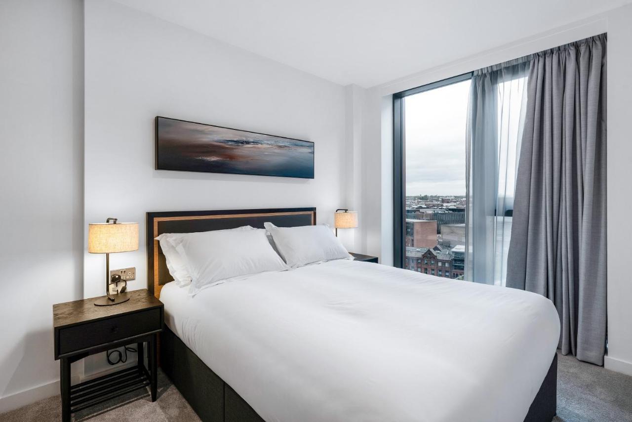 מנצ'סטר Citysuites 2 Aparthotel מראה חיצוני תמונה The image shows a modern bedroom featuring a large bed with a white duvet and two pillows. On either side of the bed are small nightstands with lamps. A large window occupies one wall, draped with light gray curtains, allowing natural light to enter 