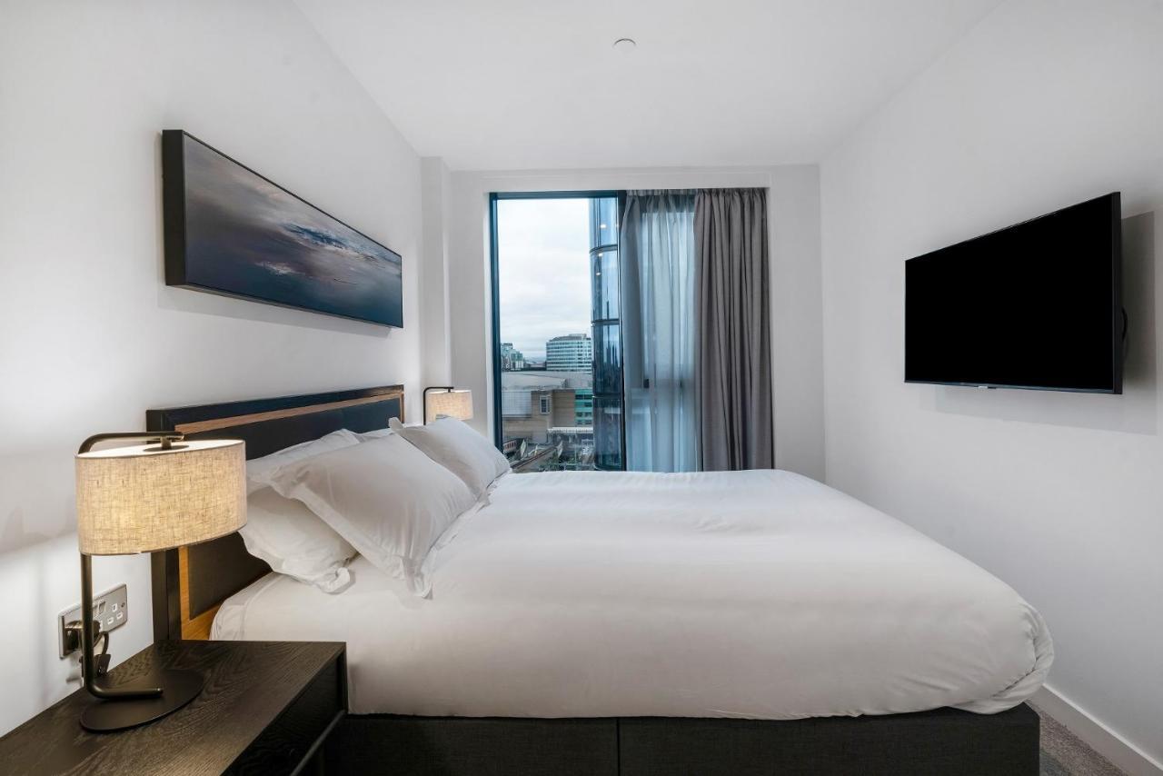 מנצ'סטר Citysuites 2 Aparthotel מראה חיצוני תמונה The photo shows a modern bedroom with a minimalist aesthetic. It features a large bed with white bedding and two pillows. On either side of the bed, there are bedside tables with lamps. A large window lets in natural light, covered by sheer curtains.
