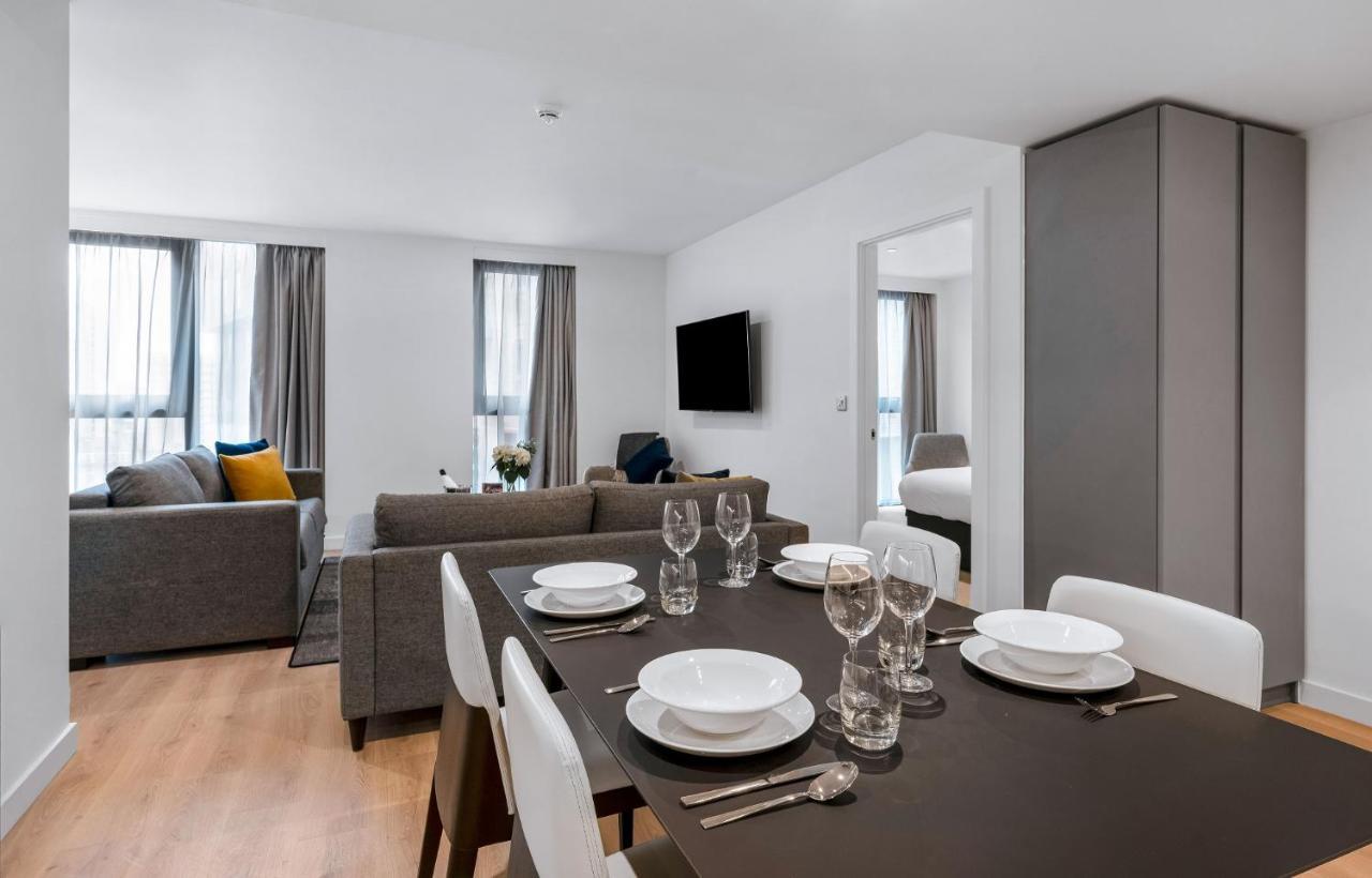 מנצ'סטר Citysuites 2 Aparthotel מראה חיצוני תמונה The photo shows a modern living space with an open layout. In the foreground, there is a dining table set for a meal, featuring multiple plates, glasses, and cutlery arranged neatly. In the background, there is a comfortable-looking gray sofa with a 