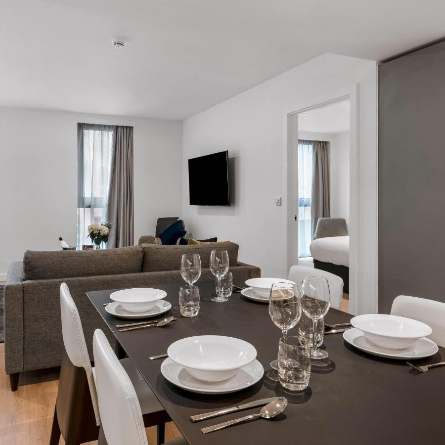 מנצ'סטר Citysuites 2 Aparthotel מראה חיצוני תמונה The photo shows a modern, well-lit living space that features a dining area and a sitting area. In the foreground, there is a dining table set for six, with white plates and crystal glasses arranged neatly. The table is surrounded by white chairs. 

