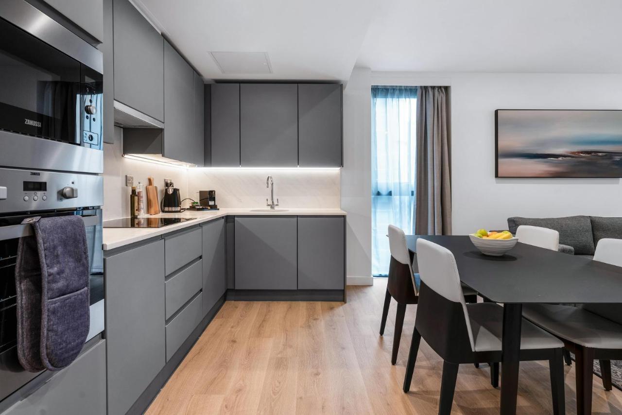 מנצ'סטר Citysuites 2 Aparthotel מראה חיצוני תמונה The photo shows a modern kitchen and dining area. The kitchen features sleek gray cabinetry, a white countertop, and built-in appliances such as an oven. There is a sink and some kitchen utensils displayed. The adjacent dining area contains a black t