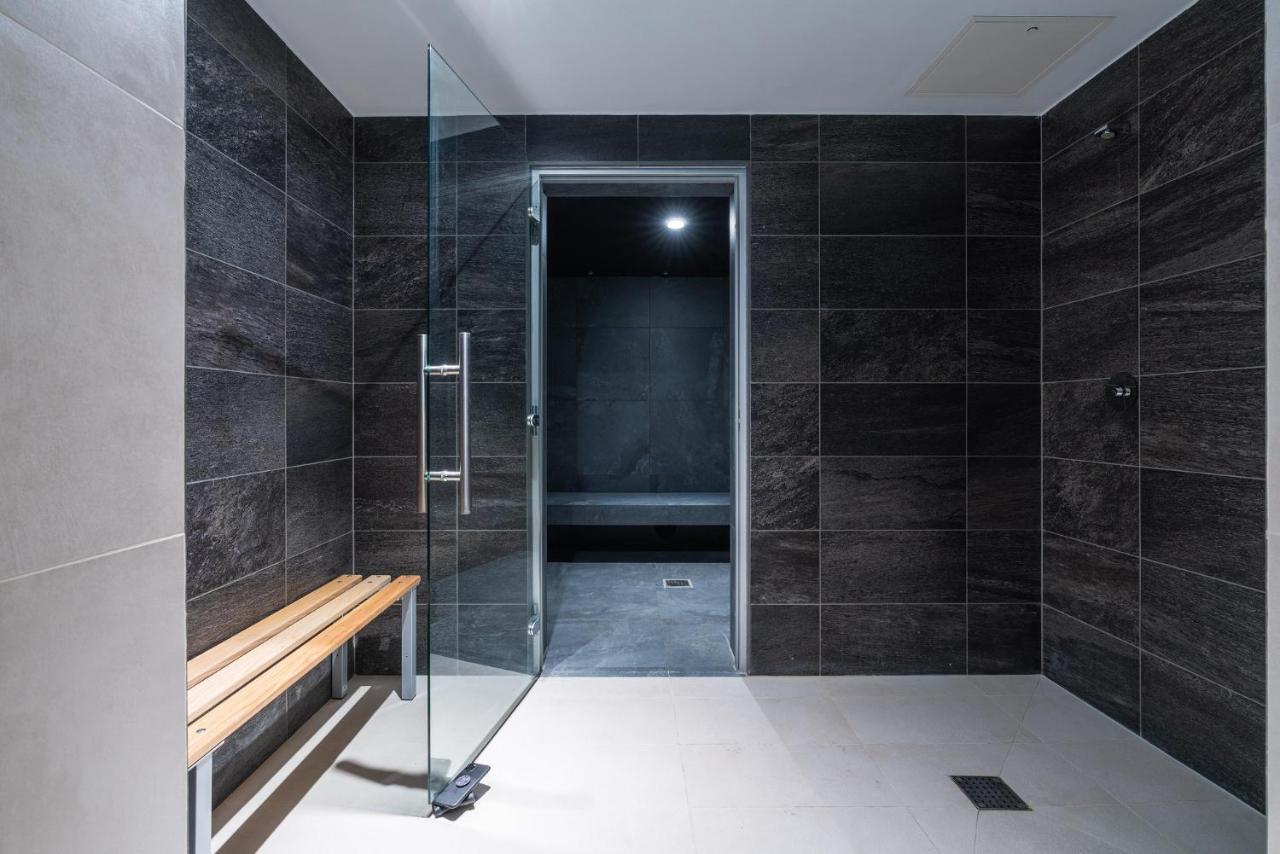 מנצ'סטר Citysuites 2 Aparthotel מראה חיצוני תמונה The photo shows a modern, minimalist shower area. The space is predominantly tiled with dark gray tiles on the walls, giving it a sleek and sophisticated look. There is a glass door that leads to another room, and a wooden bench is positioned against