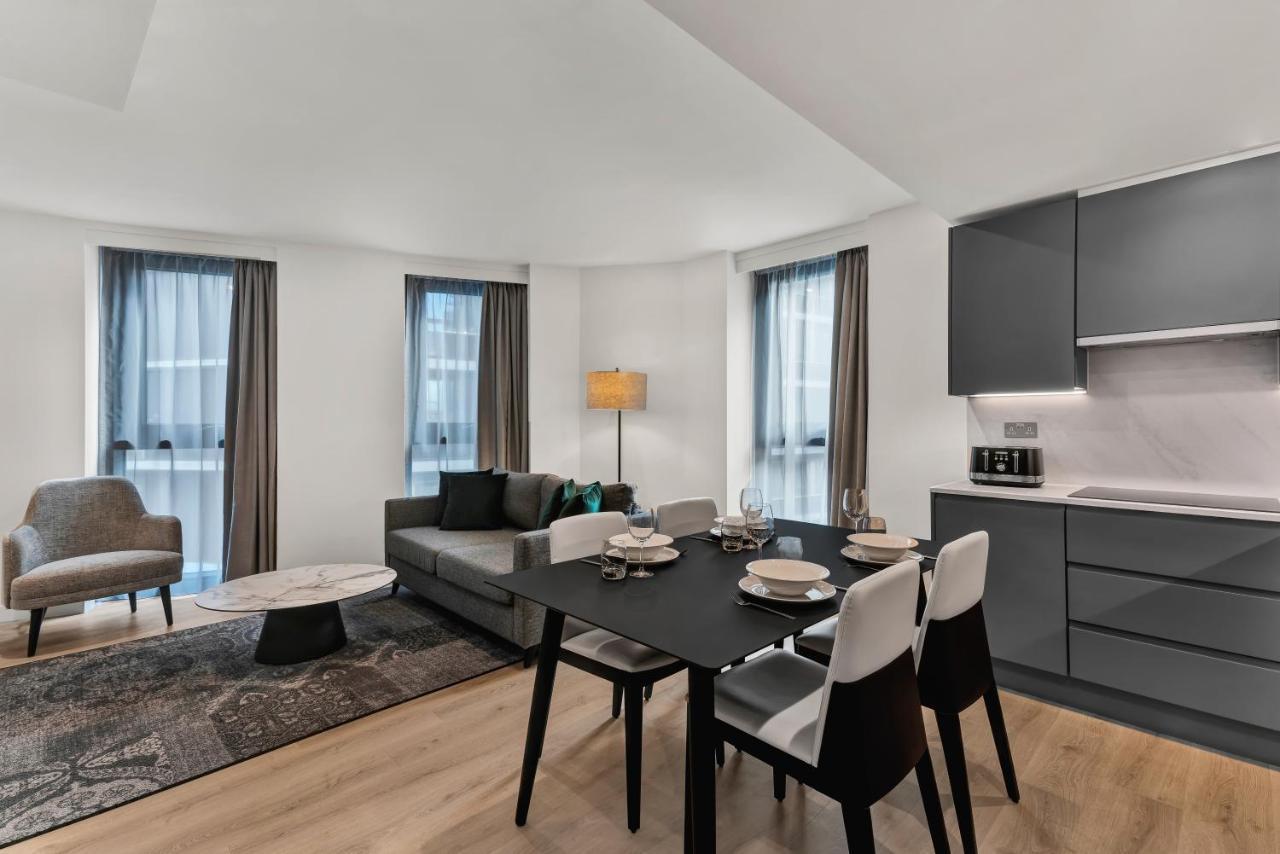 מנצ'סטר Citysuites 2 Aparthotel מראה חיצוני תמונה The photo shows a modern living space that includes a dining area and a seating area. There's a sleek black dining table set with plates and cutlery, accompanied by white and black chairs. To one side, a comfortable gray sofa faces the dining area, c