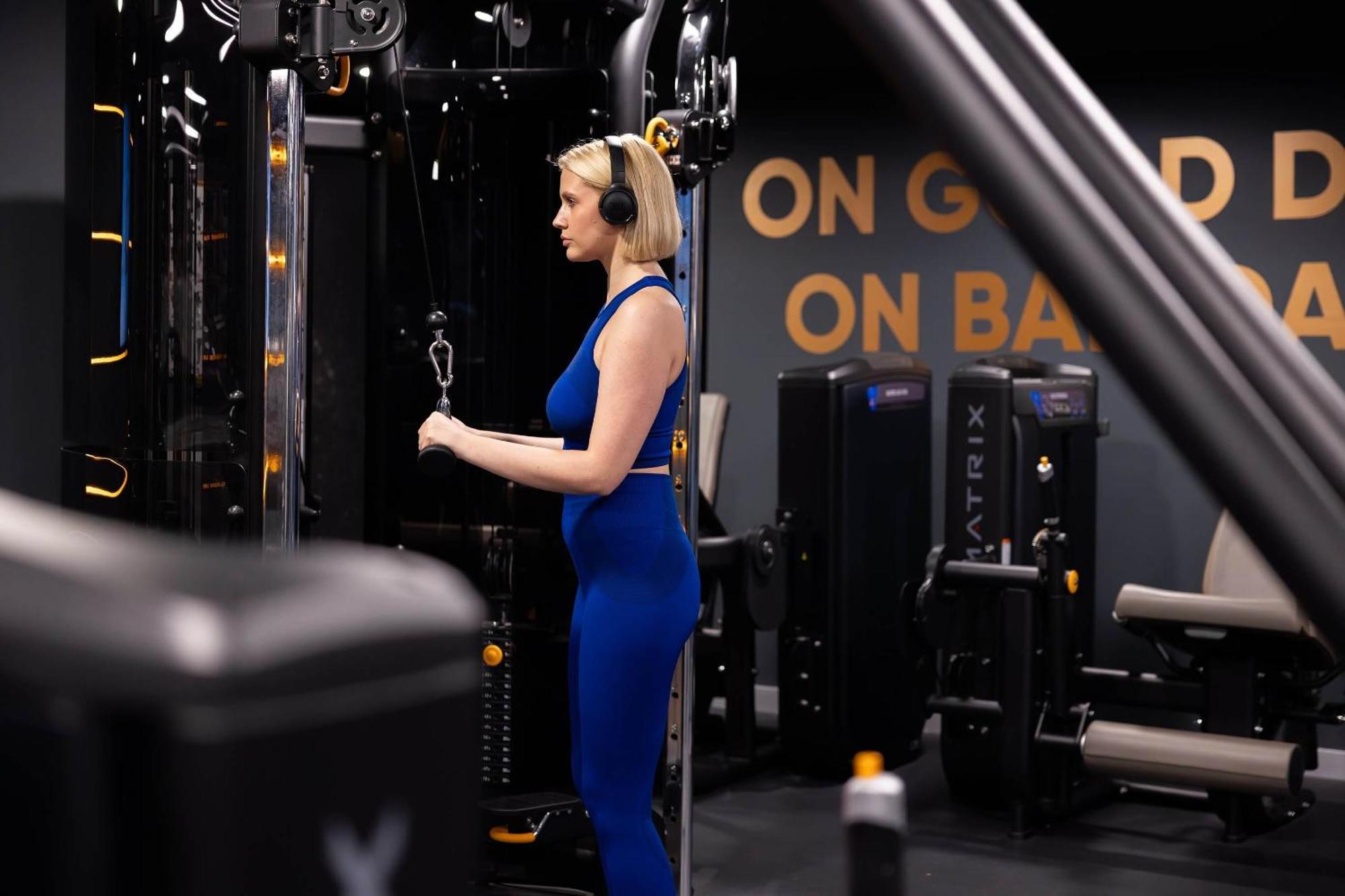 מנצ'סטר Citysuites 2 Aparthotel מראה חיצוני תמונה The photo shows a woman working out in a gym. She is wearing a blue athletic outfit and headphones while using a gym machine. The background features various other gym equipment and a wall with the words "ON GOOD ON BAD" displayed prominently. The se