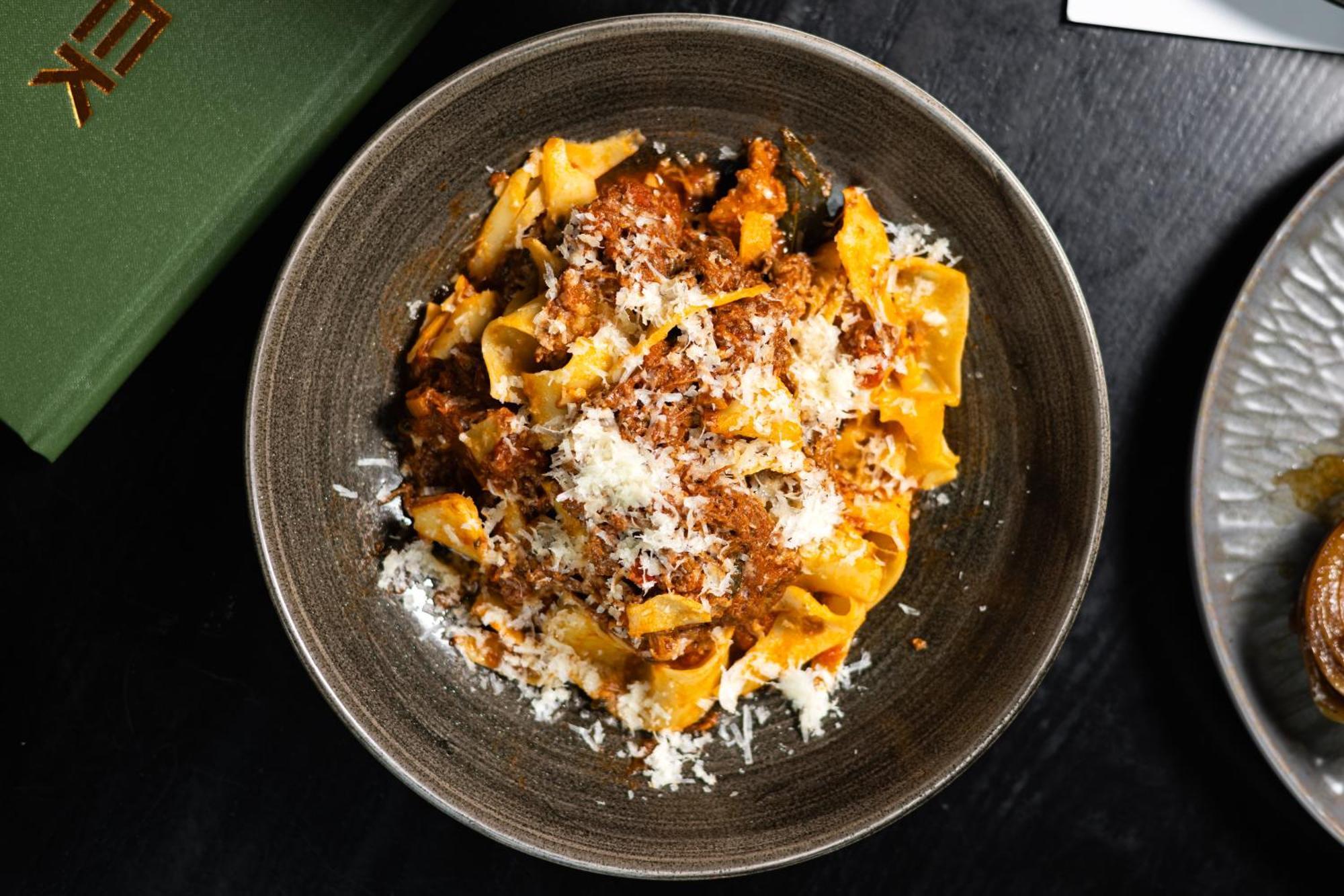 מנצ'סטר Citysuites 2 Aparthotel מראה חיצוני תמונה The photo shows a bowl of pasta featuring wide, ribbon-like noodles, likely pappardelle, fully coated in a rich meat sauce. The dish is garnished with a generous sprinkling of grated cheese, adding a creamy texture to the presentation. The dark bowl 