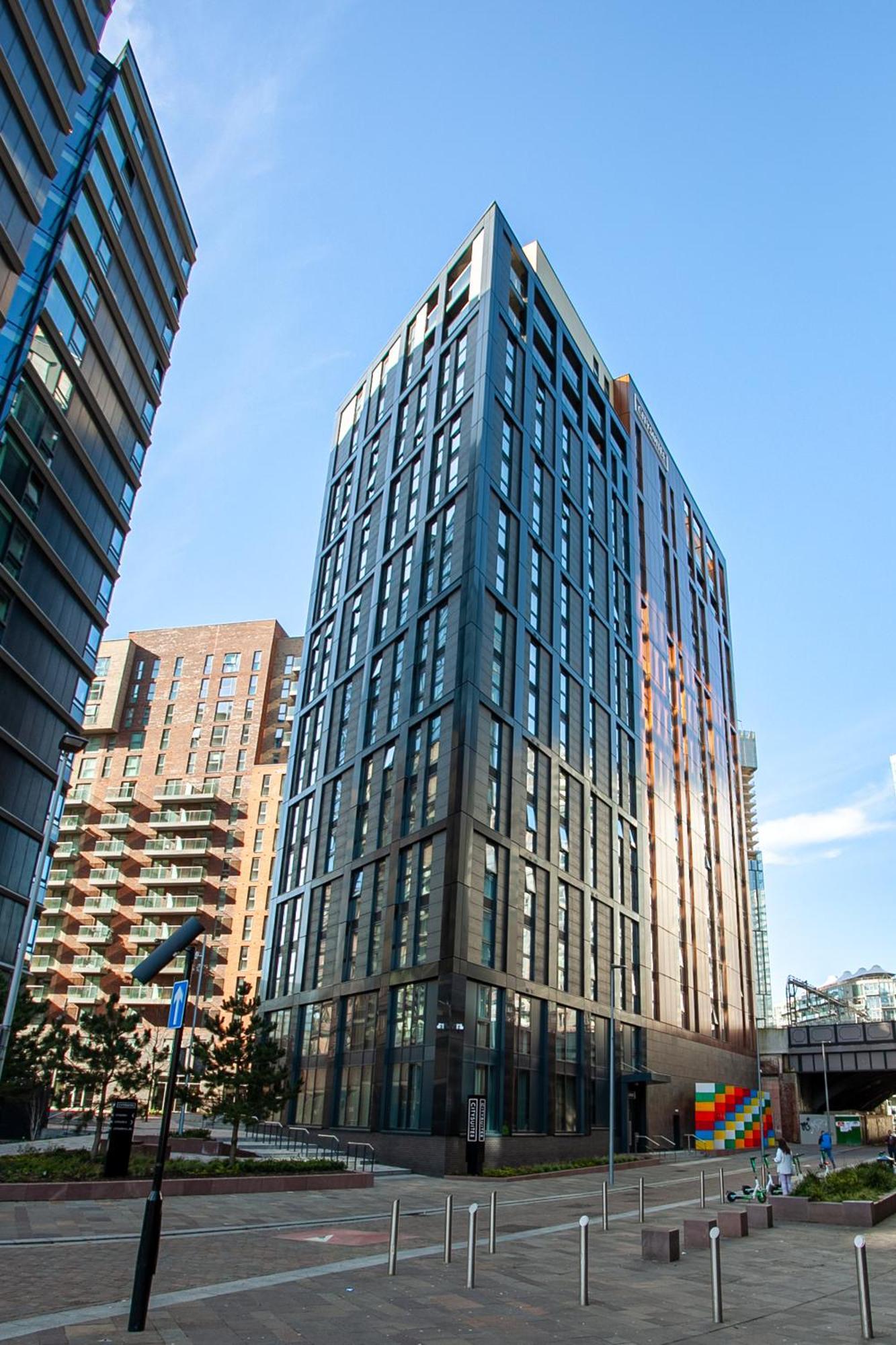 מנצ'סטר Citysuites 2 Aparthotel מראה חיצוני תמונה The photo shows a modern high-rise building with a sleek, glass façade. It is set against a blue sky, indicating a clear day. The building appears to have multiple floors with large windows that reflect light. Nearby, there are additional buildings, 